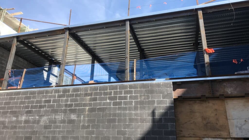Metal decking being installed at a building’s construction site. There is a large gray brick wall in front of and below the metal decking. 